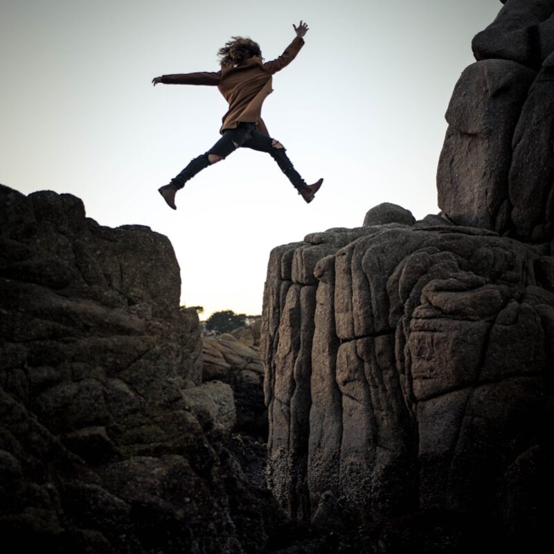 person jumping on big rock under gray and white sky during daytime