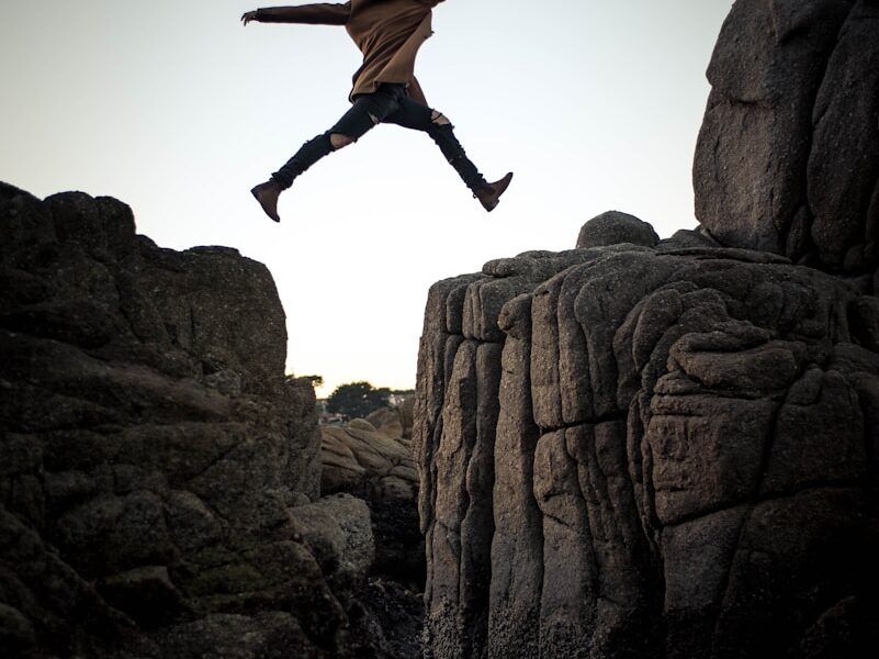 person jumping on big rock under gray and white sky during daytime